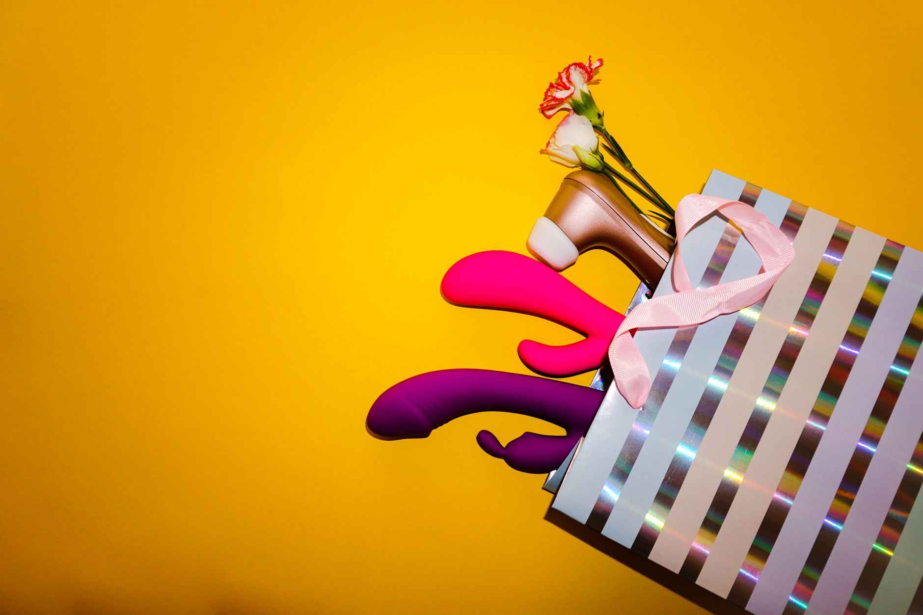 Colorful vibrators with flowers in a holographic gift bag displayed against a bold yellow background.